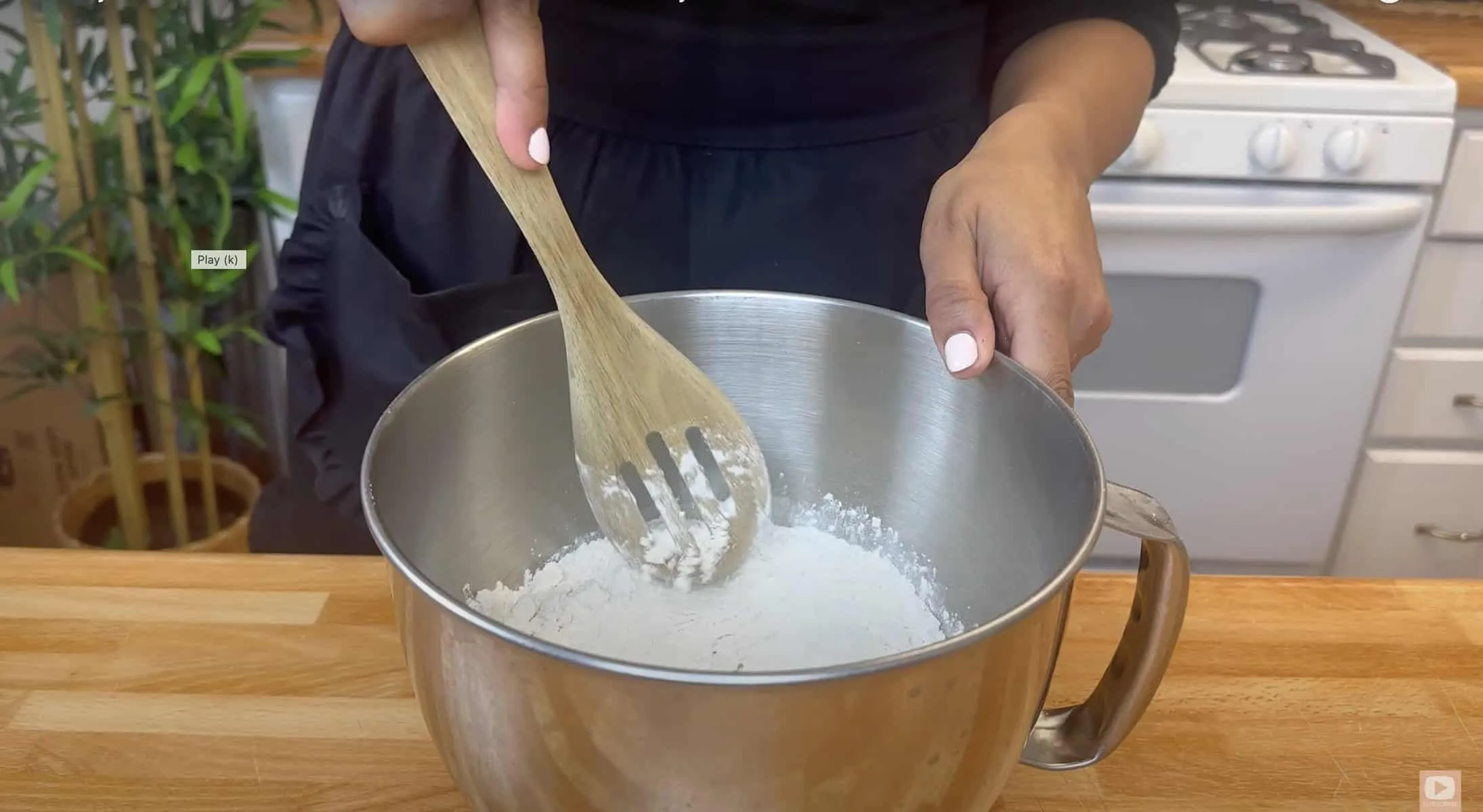 Person mixing flour in stainless bowl with wooden spoon on kitchen counter. Baking preparation scene.